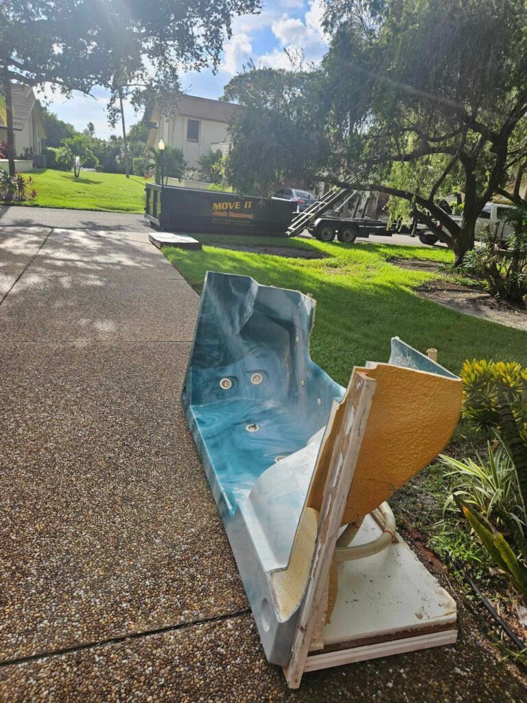 A discarded bathtub lies on a driveway beside a dumpster, with green grass and trees in the background.