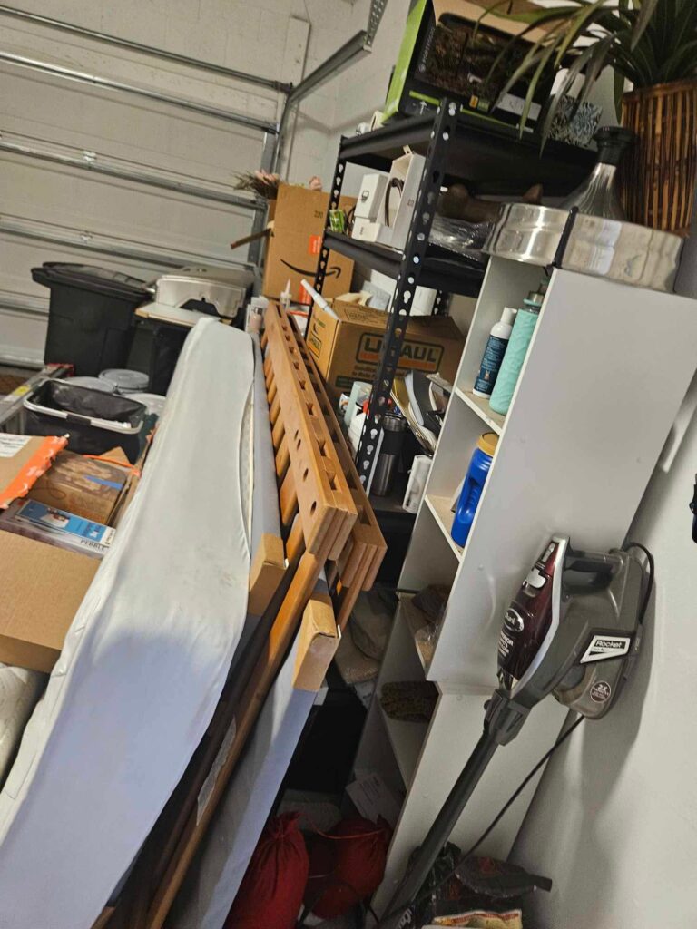 A cluttered garage space filled with boxes, shelves of items, and a bed frame.