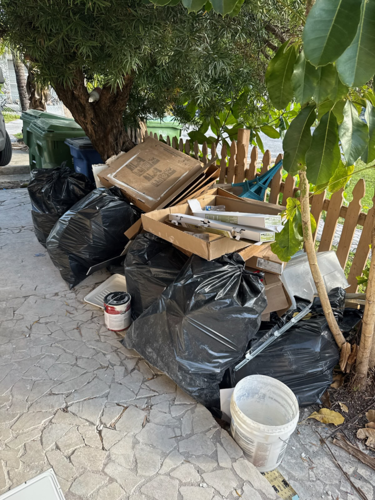 A pile of black garbage bags and cardboard boxes is stacked beside a fence, with some containers nearby.