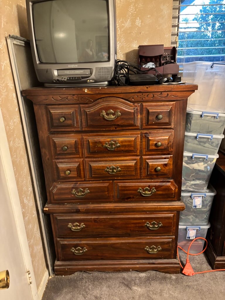 A wooden chest of drawers with multiple drawers and a small TV on top, located in a room with a window.