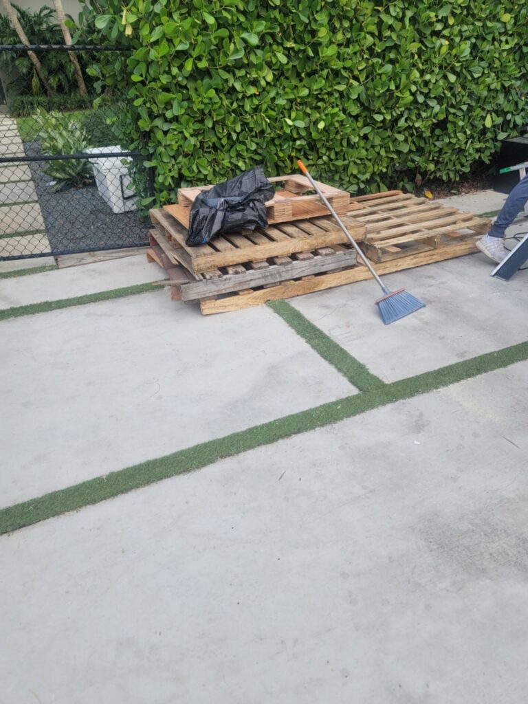 A stack of wooden pallets with a black tarp and a broom on a concrete surface, surrounded by greenery.
