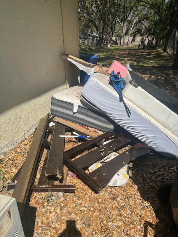 A pile of discarded furniture, including a mattress, wooden planks, and other debris, near a building.